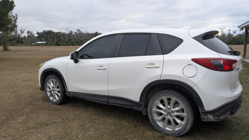 Side and rear view of a dirty, white SUV (Mazda CX-5) parked in a grassy field.