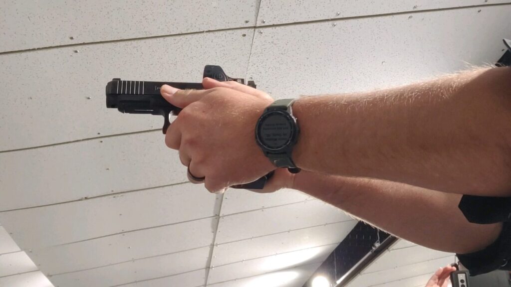 Shooter aiming a pistol with a red dot optic downrange at paper targets at a shooting range.