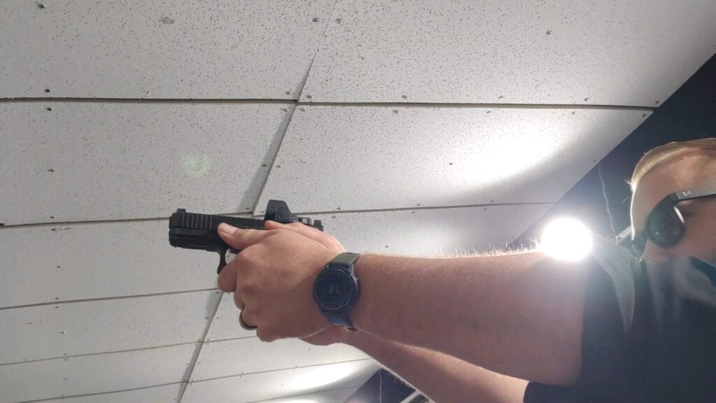 Shooter aiming a pistol with a red dot optic downrange at paper targets at a shooting range.