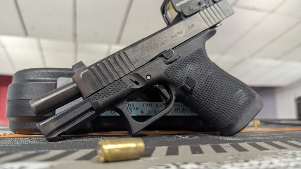 Low-angle view of a Glock 19 Gen 6 with an optic, showing the exposed barrel and a brass casing in the foreground.
