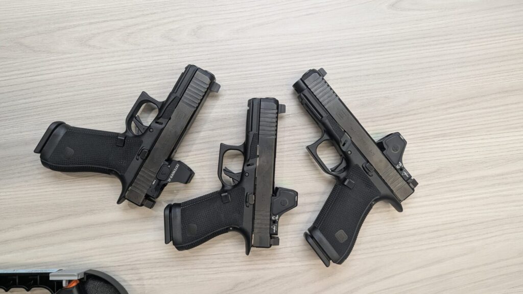 Overhead view of three black Glock Gen 6 pistols with red dot optics on a light surface,