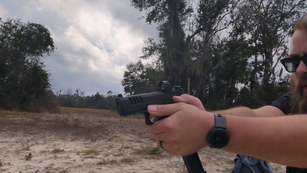 Shooter aiming a pistol with a red dot sight in a two-handed grip at an outdoor range.