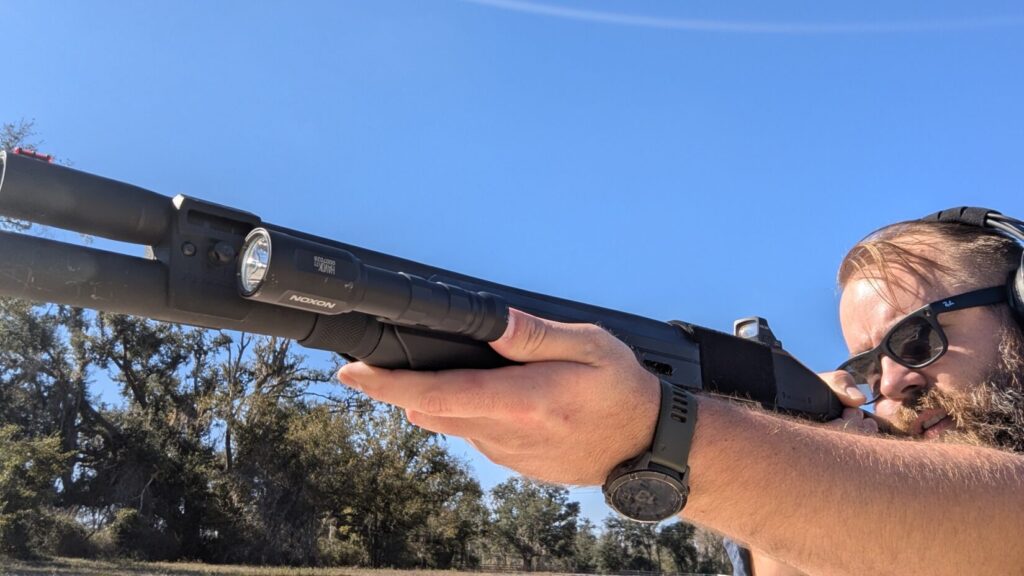 Man with beard and sunglasses holding a shotgun outdoors, looking up, with a Modlite Havok mounted.