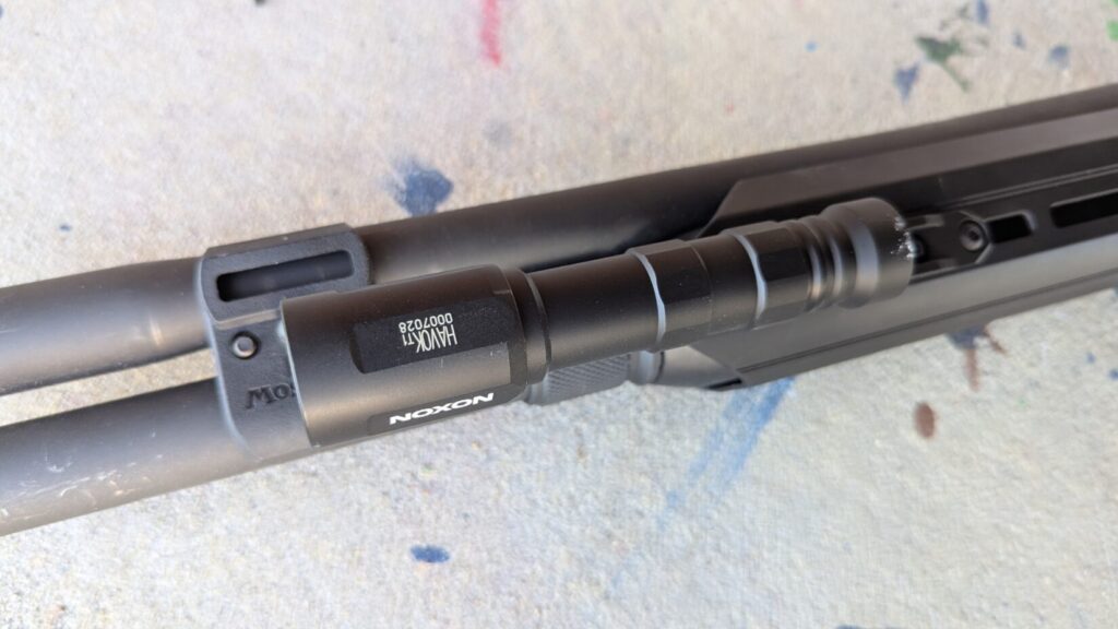 Close-up of the Modlite Havok flashlight mounted on the shotgun's magazine tube, showing "HAWOK" and serial number.