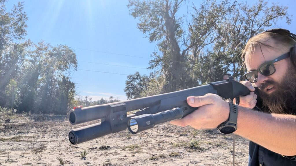 Man with beard and sunglasses holding a shotgun outdoors, looking up, with a Modlite Havok mounted.