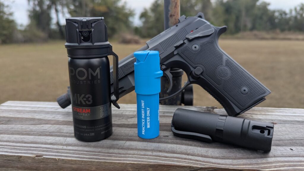 Three POM pepper spray canisters (MK-3, blue inert practice unit, and a small black unit) with a handgun in the background.