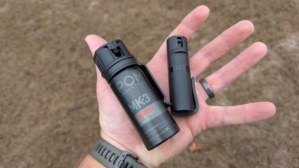 Hand holding a black POM pepper spray canister, with the thumb placed near the flip-top safety.