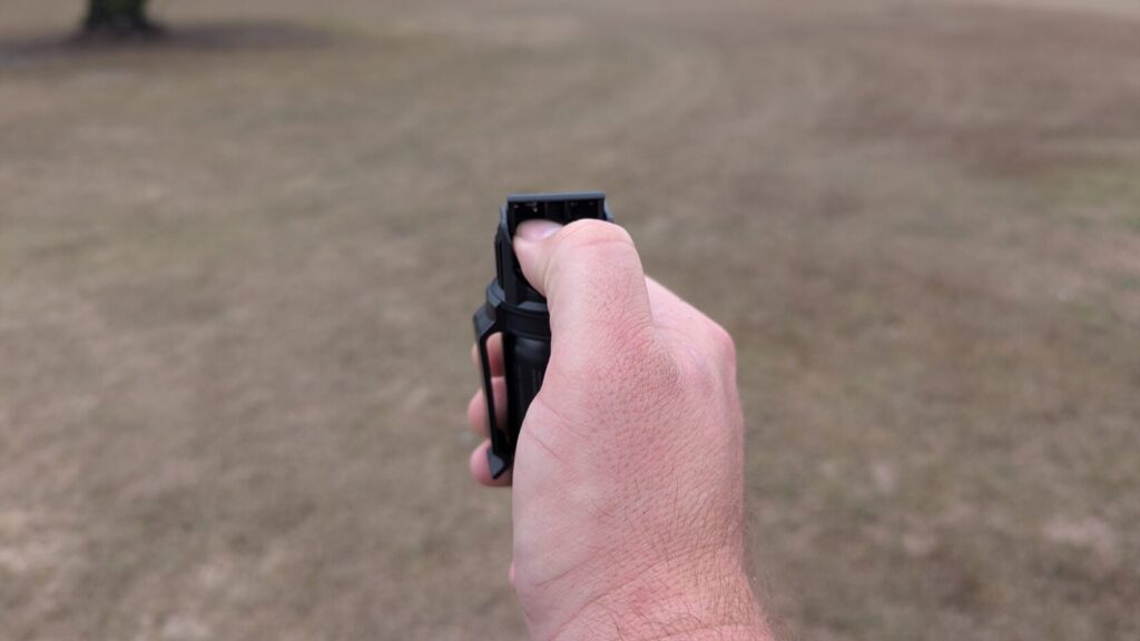 Hand pressing down the actuator button on a black POM pepper spray unit after lifting the safety.