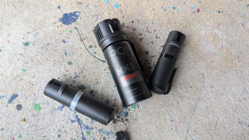 Three black POM pepper spray units of different styles lying on a concrete surface.