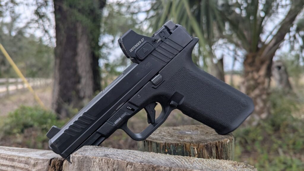 Black Ruger RXM pistol with a red dot sight balanced on a wooden post and rail outdoors.