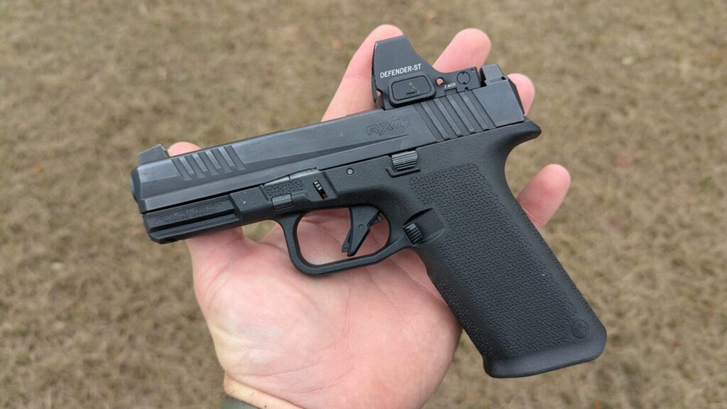 Black Ruger RXM pistol with a Defender-ST red dot sight held in the palm of a hand over a grassy background.