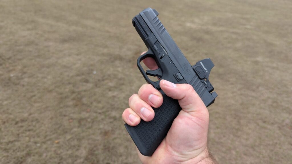Hand holding a black pistol with a red dot sight outdoors.