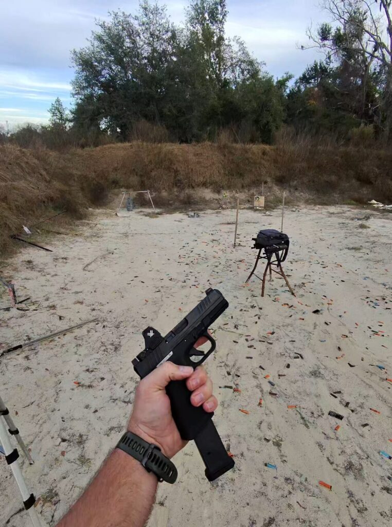 Hand holding a black pistol with a red dot sight outdoors.