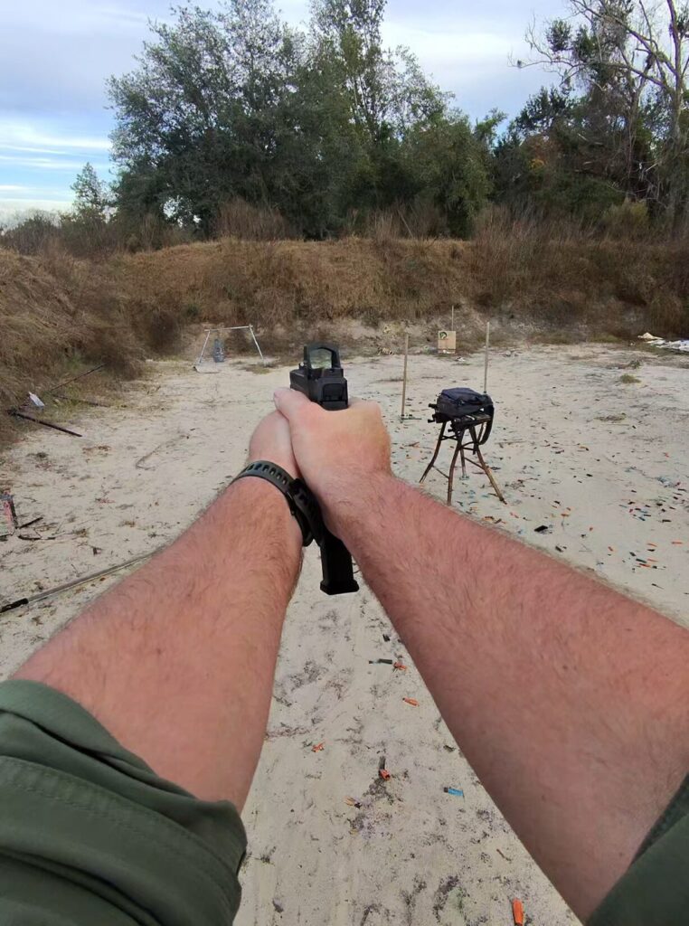 Shooter holding a Ruger RXM pistol with an extended magazine and red dot sight on an outdoor sand range.