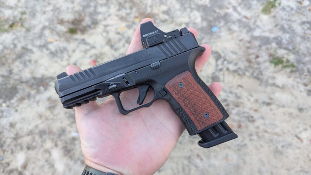 Hand holding a black pistol with a Sharps Bros grip module, wood grip panels, and a red dot optic.