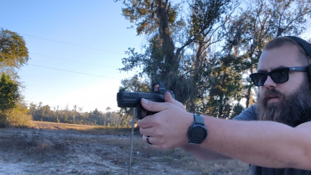 Man aiming a pistol outdoors, showing his watch and shooting stance.