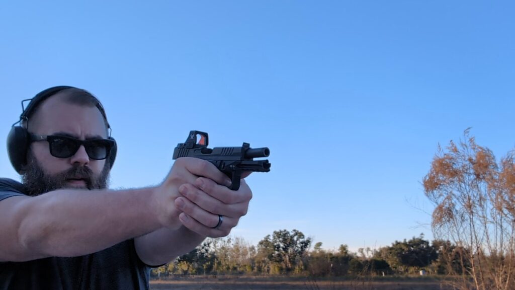 Man shooting a pistol outdoors right after firing, with muzzle blast visible.