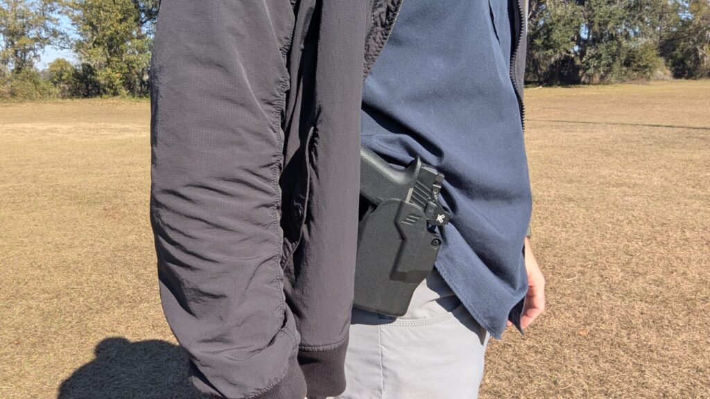 Man standing outdoors with a handgun holstered on his hip, mostly concealed by a black jacket.