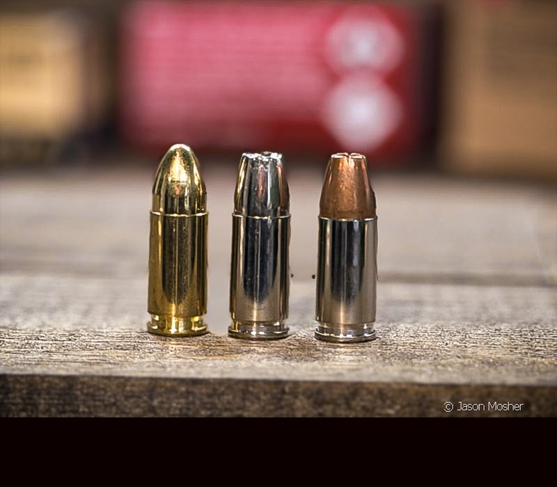 Three 9mm cartridges standing on their back, in front of boxes of ammo. 