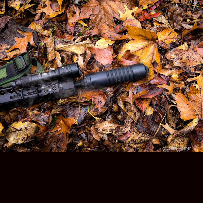 A black A-10 silencer attached to an AR-15, laying in the leaves. 