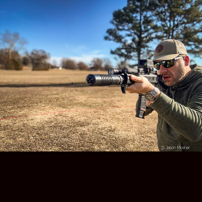 A person in a green hoody and green hat, shooting a black rifle with a black A-10 silencer attached. 