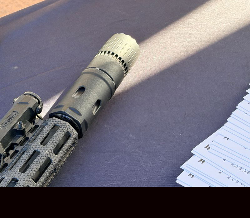 A tan rifle with a tan silencer attached, laying on a black table. 