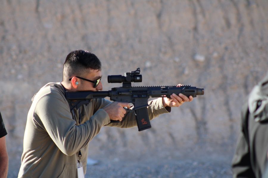 A man in a brown shirt, shooting a black rifle on the range. 