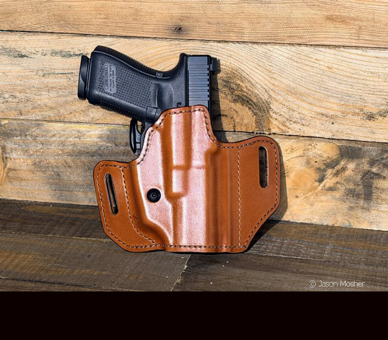 A brown leather holster that has a black gun in it, leaning up against a wood wall.
