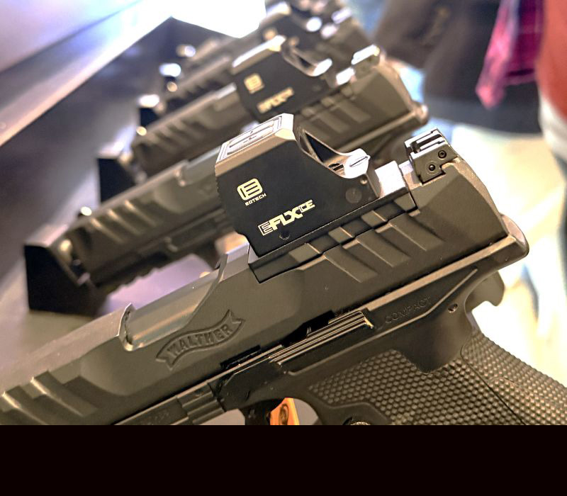 A view of the side of a gun with an optic mounted on top and several more handguns lined up behind it.