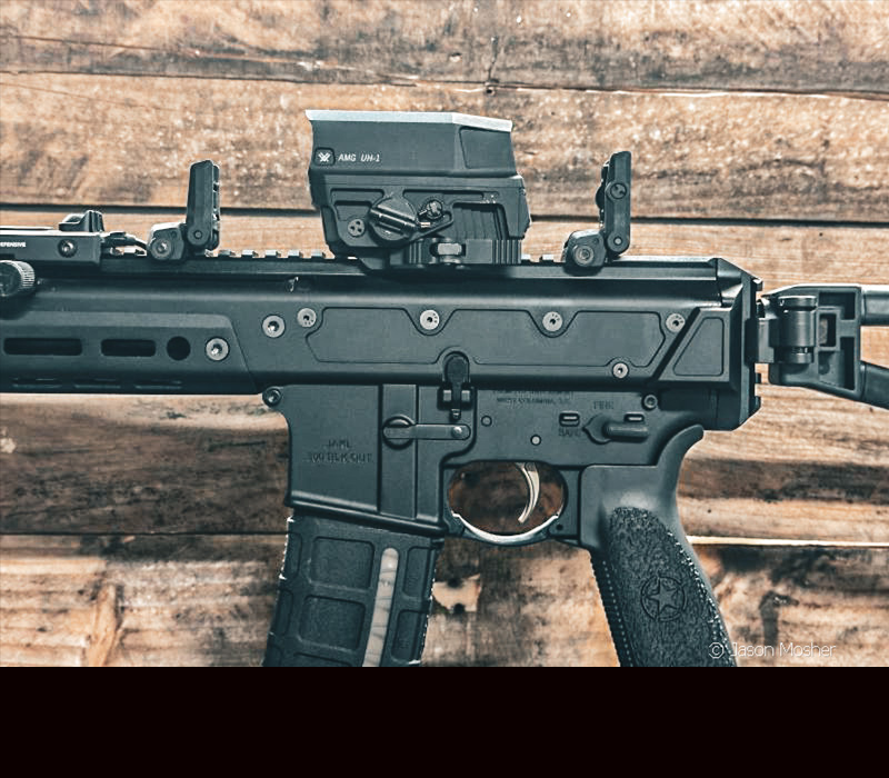 A black JAKL firearm with a large optic and two black flip-up sights in front of a wooden wall. 