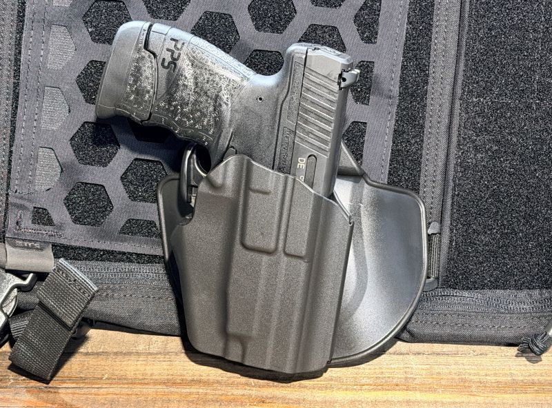 A black PPS M2 in a black Safariland holster in front of a black bag on a wood table.