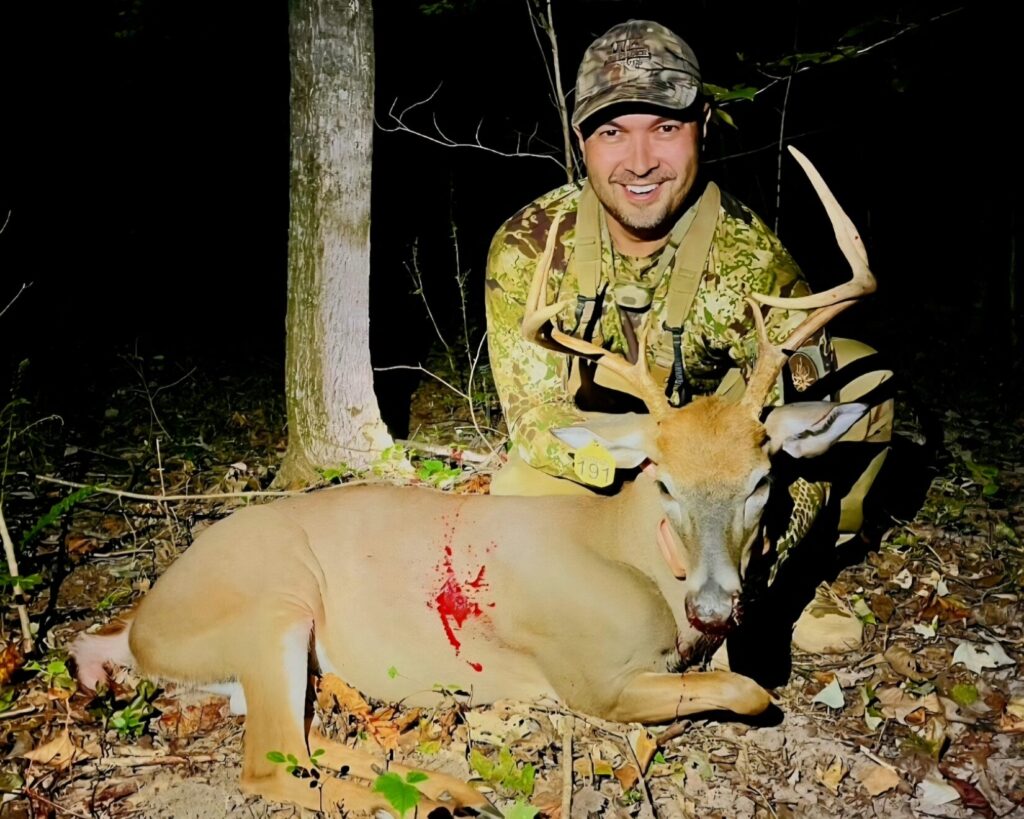 A man in camouflage next to a whitetail buck in dark woods