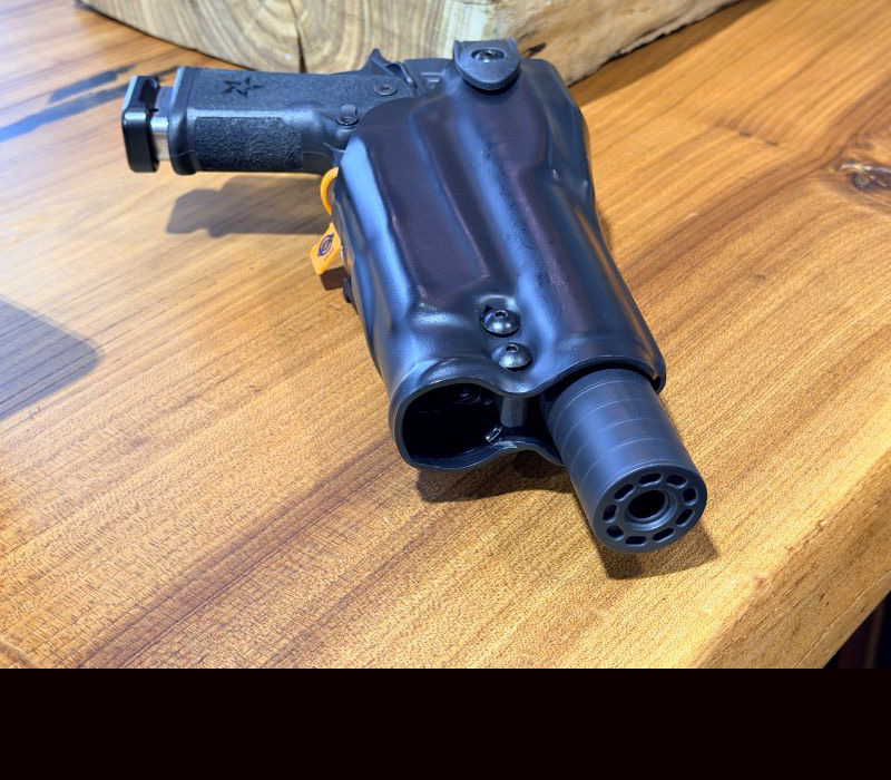 A black holster on a wood counter, with a gun in the holster that has a silencer attached to it. The silencer is sticking out of the end of the holster. 