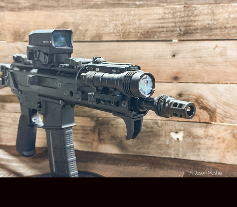 A black rifle with a light attached to the end, in front of a wooden wall. 