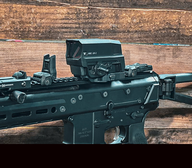 A large optic on a black rifle in front of a wood wall. 