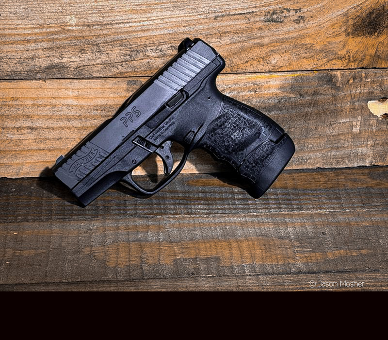 A black Walther PPS M2 handgun leaning against a wood background.