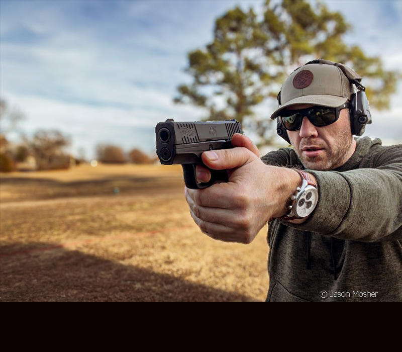 A person in a green shirt, hat, and black sunglasses, shooting the PPS M2 handgun.