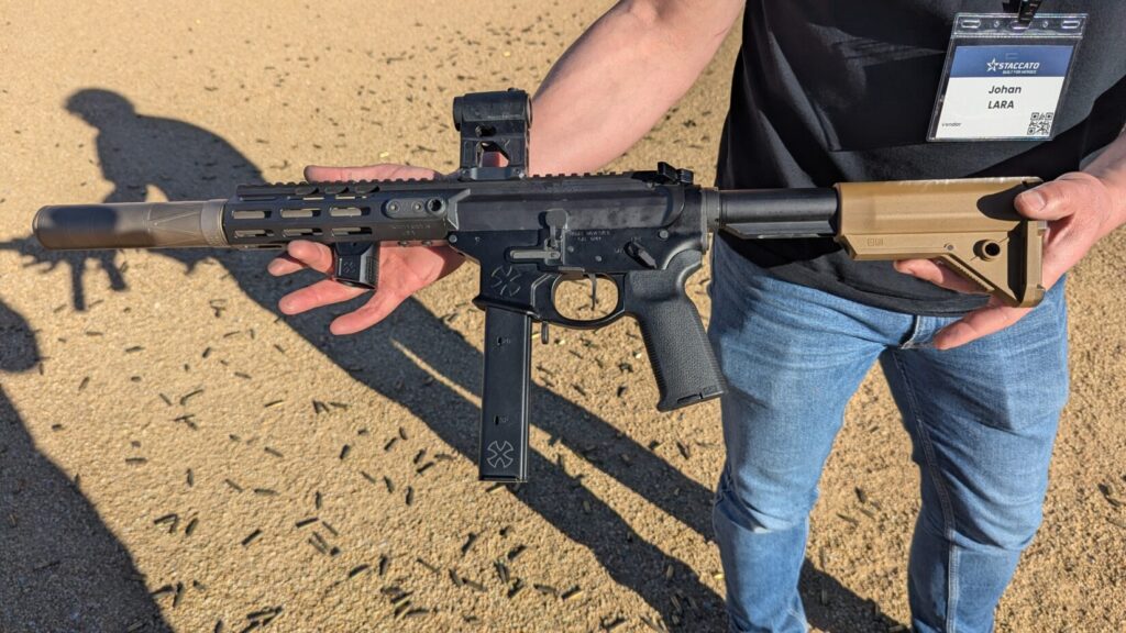 A person holding a short-barreled AR-15 style rifle equipped with a suppressor, red dot sight, and tan stock at an outdoor range.