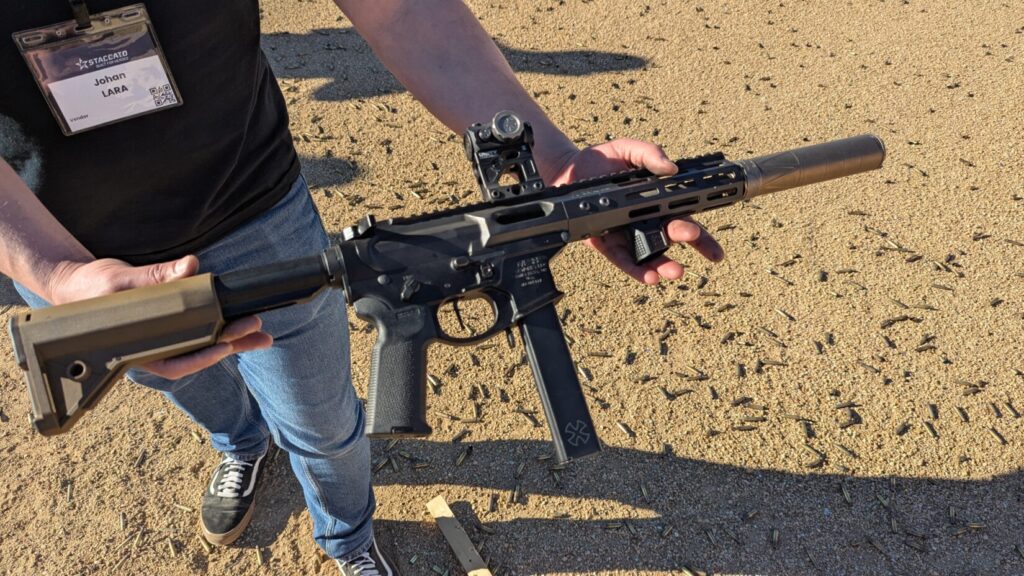 A person holding a short-barreled AR-15 style rifle equipped with a suppressor, red dot sight, and tan stock at an outdoor range.