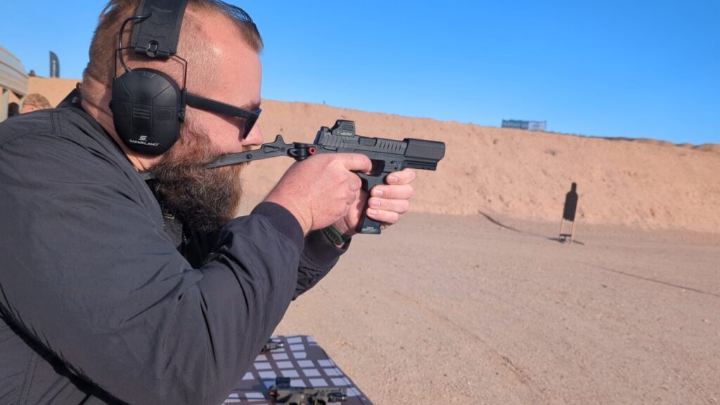 A person wearing Safariland hearing protection and sunglasses shooting a black Flux Defense Raider chassis equipped with a Holosun red dot sight at an outdoor range.