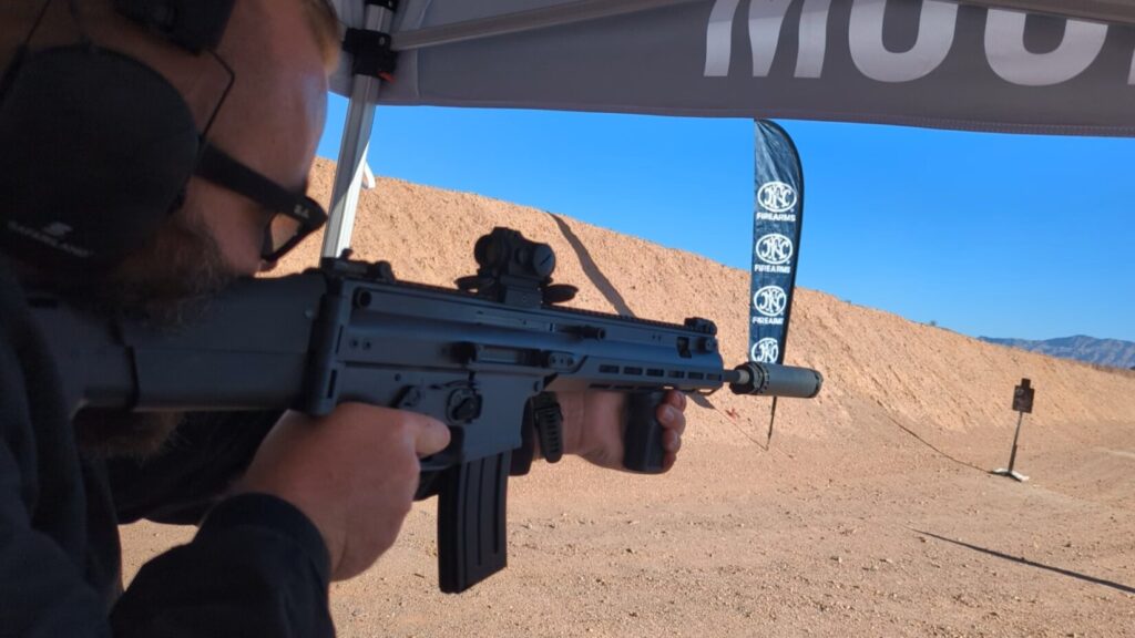 A person firing an FN SCAR rifle equipped with a suppressor and red dot sight at an outdoor shooting range.