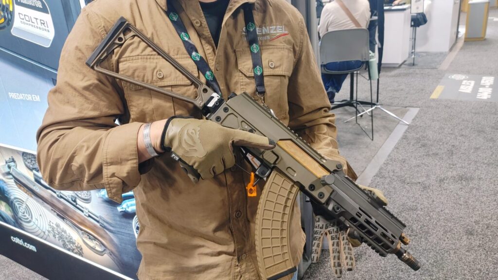 A person wearing tactical gloves holds a tan AK-style rifle featuring a folding stock and a long, curved magazine.