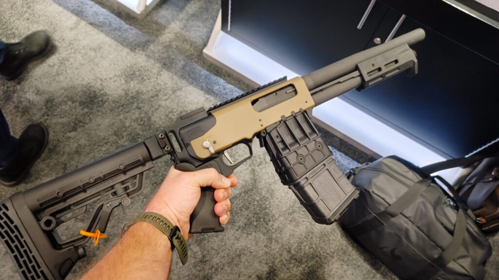 A person holds a tan Mossberg 590M magazine-fed shotgun featuring a black adjustable stock and a large detachable box magazine.