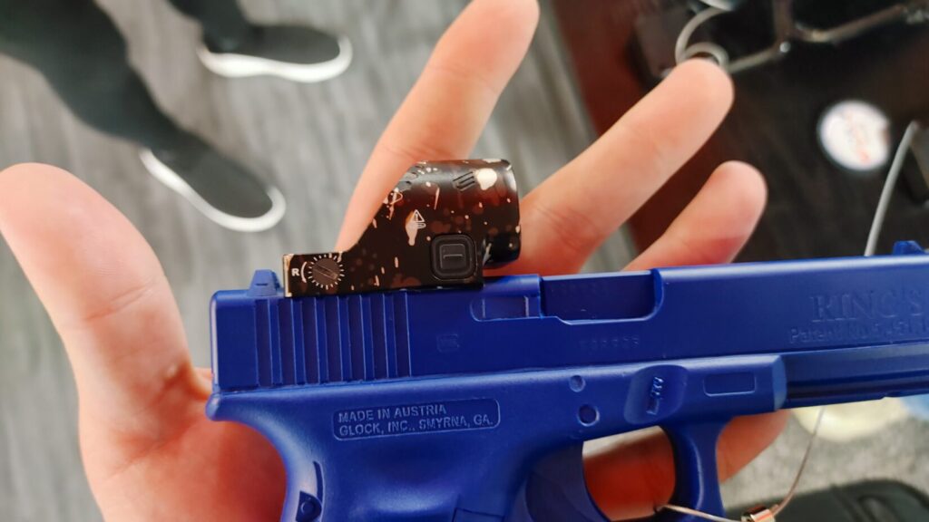 A blue training pistol held in a hand, equipped with a red dot sight featuring a brown and tan splatter camouflage pattern.