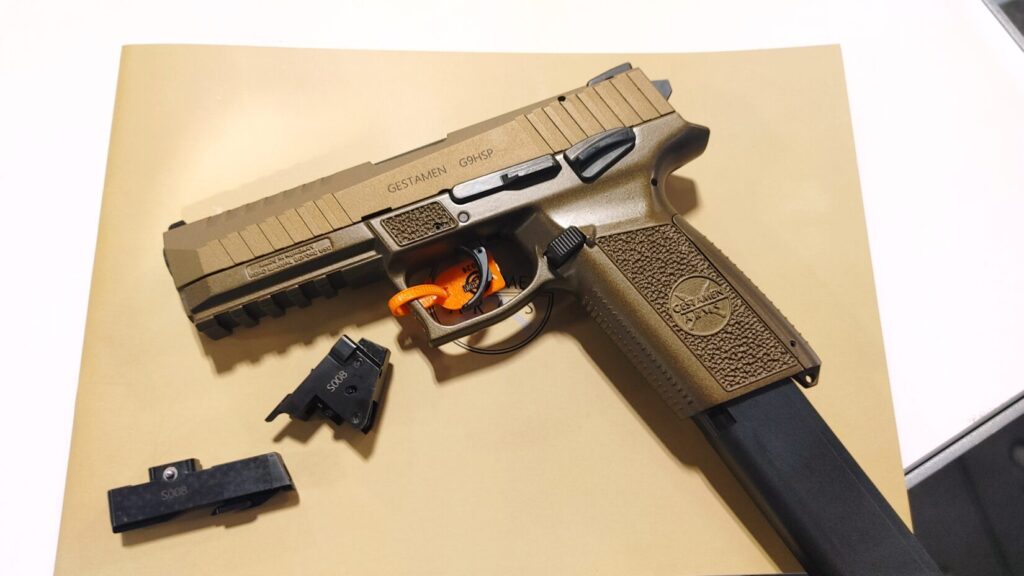 A tan Gestamen Arms G9HSP semi-automatic pistol with an extended magazine and disassembled parts on a table