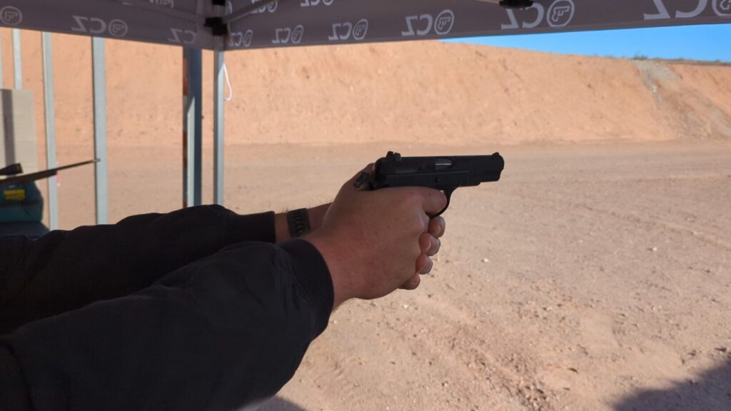 A person holding a black handgun at a range, seen from behind their hands under a branded tent.