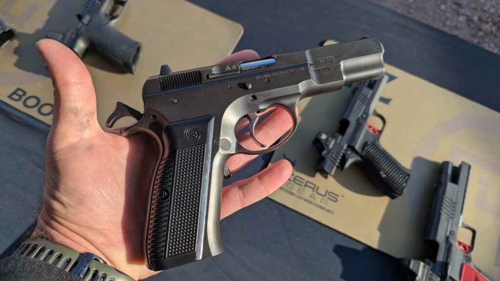 A hand holding a dark gray semi-automatic pistol with a textured black grip, with other firearms visible in the background.