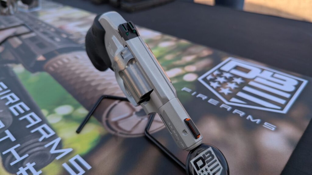 A top-down perspective of a silver revolver on a display stand, highlighting its green rear and orange front fiber optic sights.