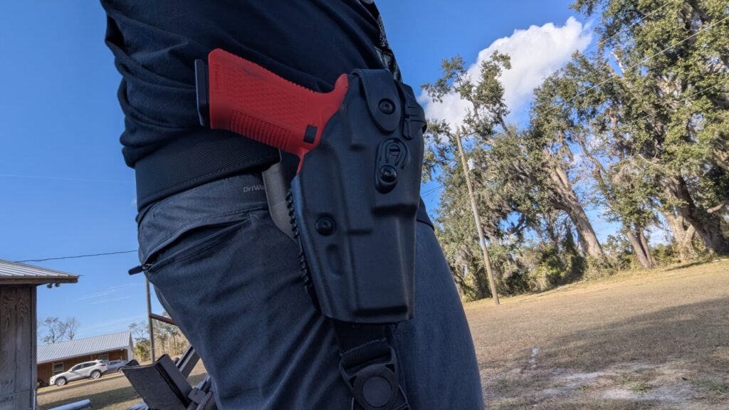 A full-length side profile of a bearded man standing in an open field, wearing a tactical belt with a red training pistol in a leg holster.