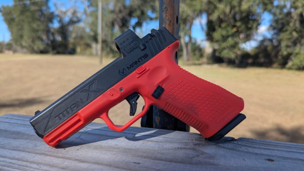A profile shot of a Mantis Titan X training pistol with a bright red frame and black slide, resting on a wooden railing.